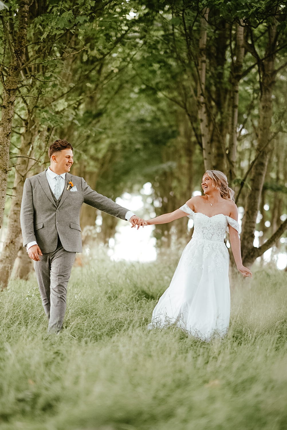 Bride and groom walking hand in hand through a woodland path at Vallum Weddings, with lush greenery and romantic natural lighting—capturing a serene countryside wedding moment in Northumberland.