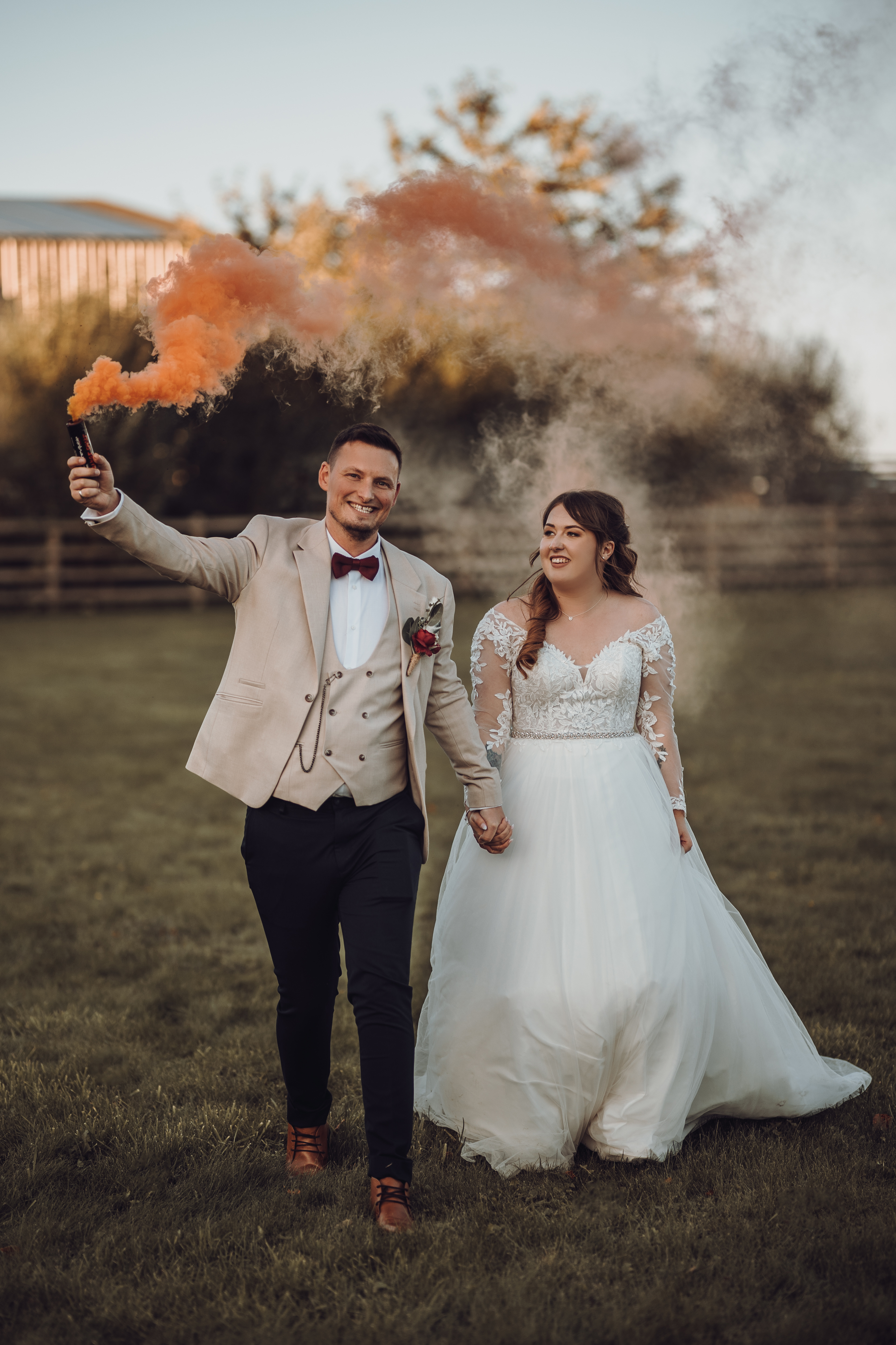 Bride and groom stroll through a countryside field at a rustic barn wedding at Vallum Weddings, with the groom waving an orange smoke bomb; the bride wears a vintage lace gown and the groom is in a beige suit and red bow tie, capturing a romantic rural moment.